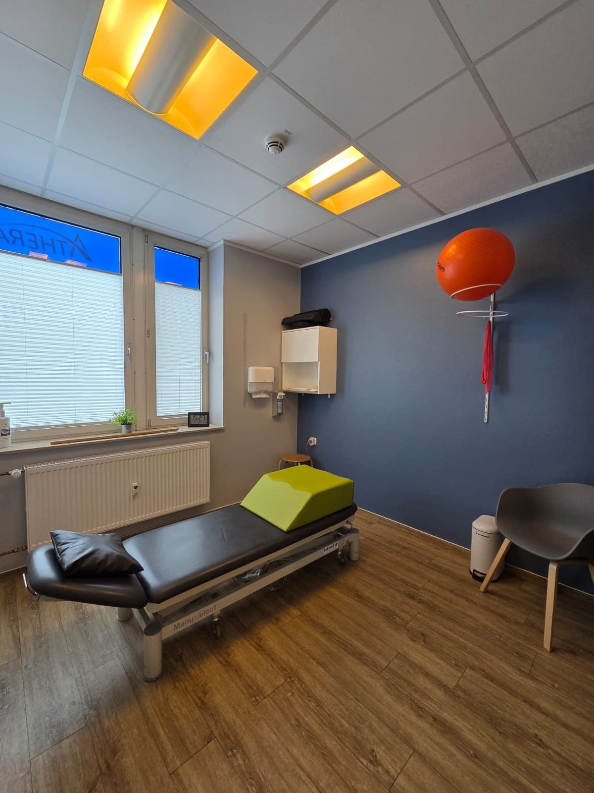 Heller Behandlungsraum mit Therapieliege, Gymnastikball und blauer Akzentwand